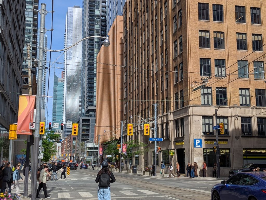 View south along Peter Street at Adelaide Street West. The Commodore building fills the right side of the image, with 30 Widmer Street’s long ribbed façade extending behind it; pedestrians cross the intersection below.